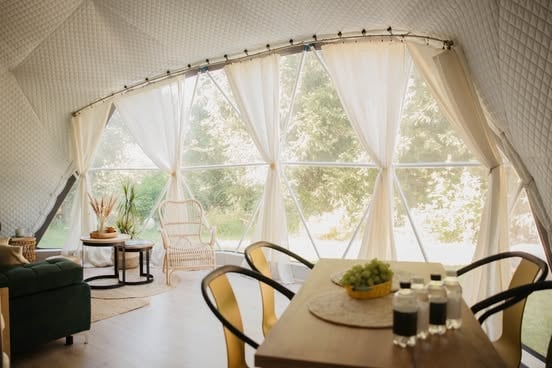 Bright dining room with curved curtain rods, sheer cream curtains over large windows overlooking trees, wooden table with black chairs, and plant décor.