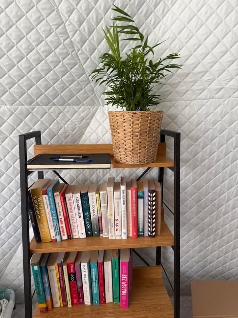 Black metal bookshelf with wooden shelves against white quilted wall, displaying books and a potted plant in woven basket on top.