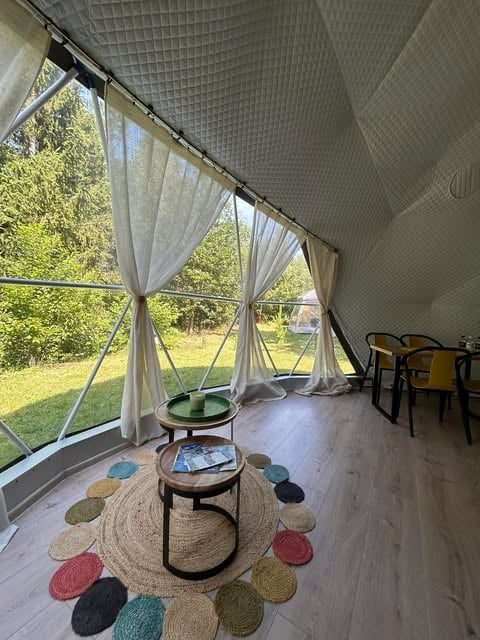 Bright screened porch interior with sheer curtains, round wooden tables, colorful circular rug, and green lawn view through windows