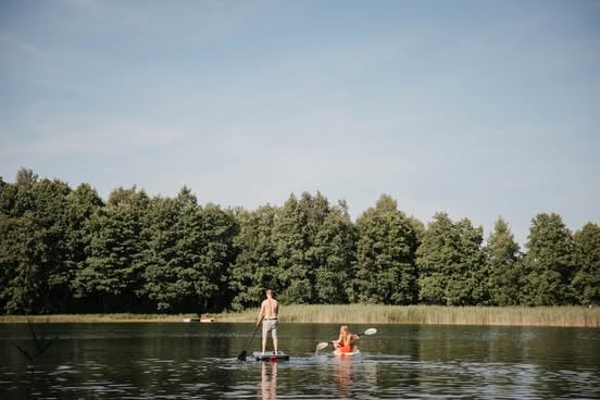 Two people paddleboarding and swimming in a calm lake with a tree-lined shoreline under a clear blue sky