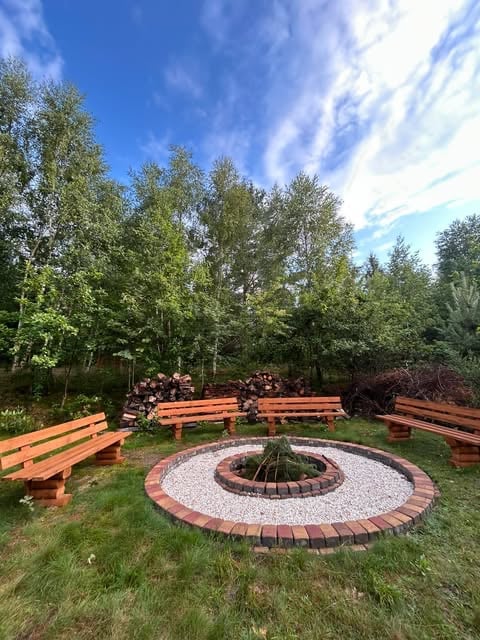 Outdoor fire pit surrounded by orange wooden benches, gravel circle, and green trees under blue sky