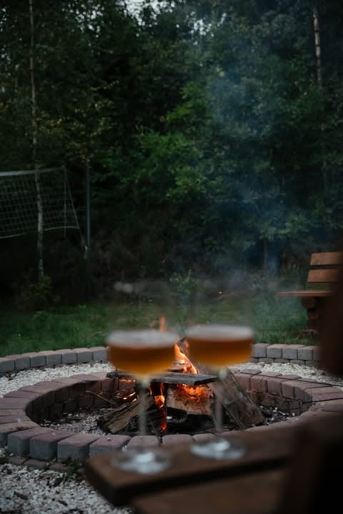 Two glasses of beer steaming over an outdoor fire pit surrounded by stone, with green forest backdrop