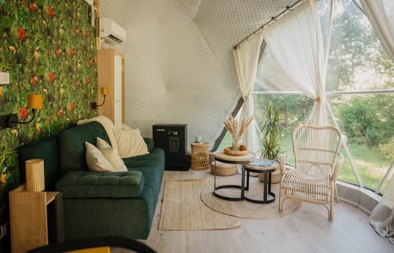 Modern living room with green armchair, botanical wallpaper, hanging rattan chair, large windows with natural light, and wooden accents