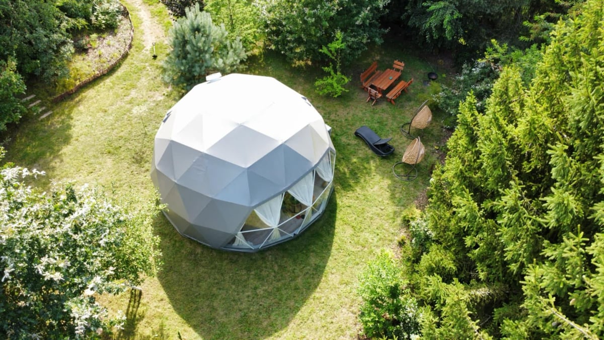 Aerial view of a white geodesic dome structure surrounded by green lawn and forest trees