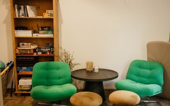 A modern living room with two bright green armchairs facing a black round table, wooden ottoman, and bookshelf against the wall