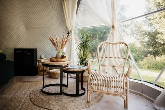 Bright sunroom with woven rattan chair, round nesting tables, potted plant, and sheer white curtains overlooking greenery