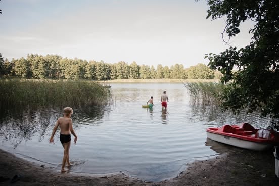 Children wading in a calm lake surrounded by reeds and trees with a red boat anchored nearby