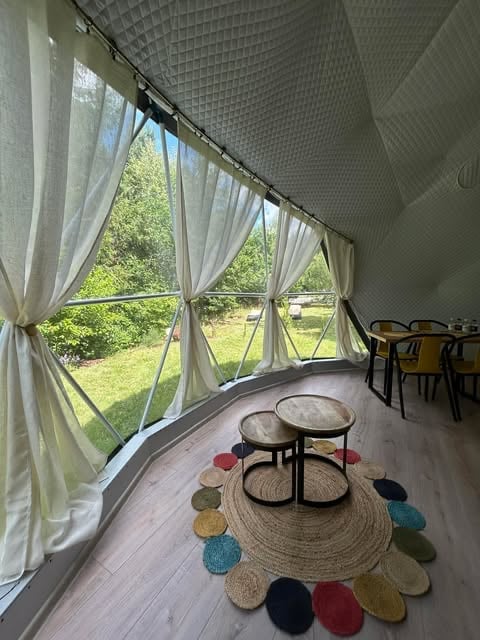 Bright sunroom with large windows, white curtains, wooden flooring, two small round tables on colorful circular rugs, and dining chairs in background