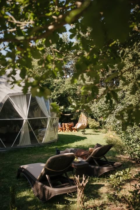 Outdoor garden lounge with black woven chairs, geodesic dome tent, and lush green foliage overhead on sunny day
