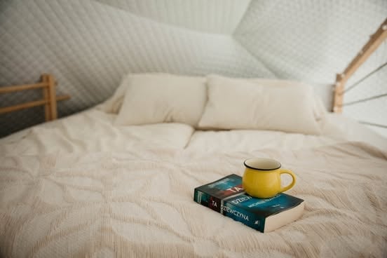 A yellow mug and book resting on a cream-colored bed in a bright attic bedroom