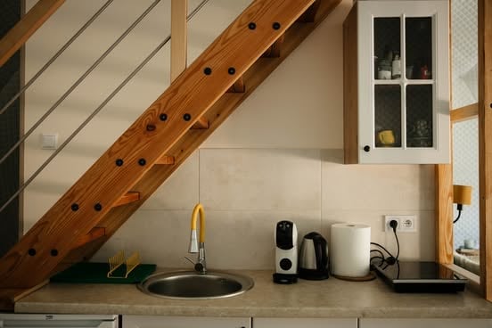 Modern kitchen counter under wooden stairs with sink, kettle, coffee maker, and storage items on beige countertop
