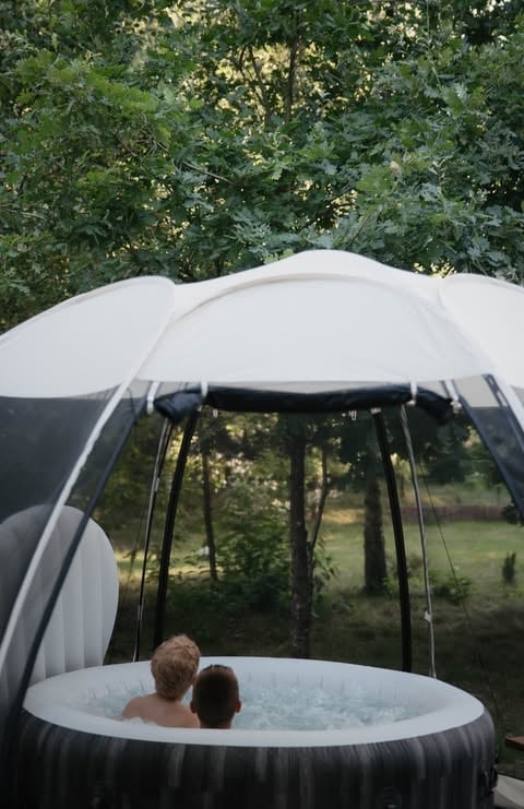 Two children enjoying an inflatable hot tub with white dome cover in a backyard surrounded by trees