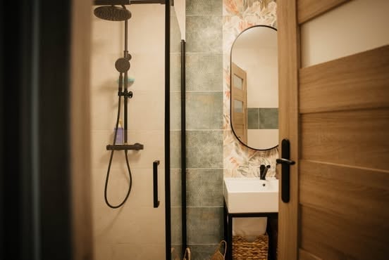 Modern bathroom with walk-in shower featuring dark metal fixtures, tiled wall, and adjoining sink vanity with oval mirror