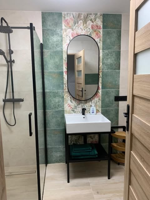 Modern bathroom with oval mirror, white sink, black metal vanity, green floral tile backsplash, and rainfall shower system with black fixtures